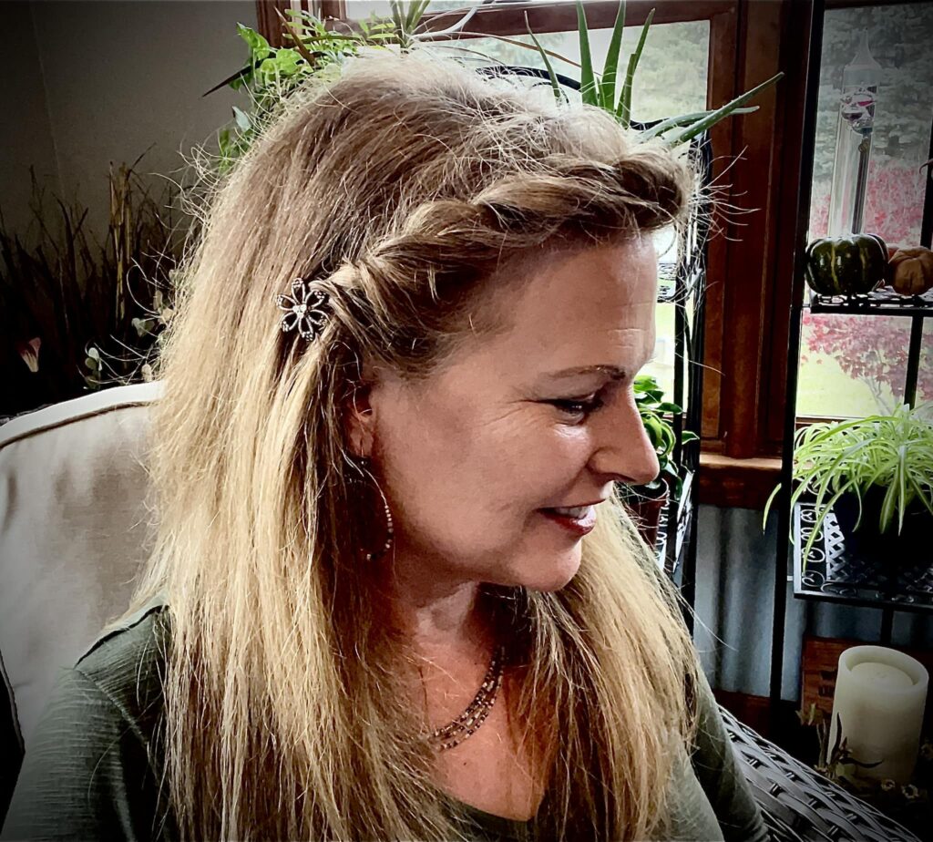 Middle age woman sitting in a soft tan chair siting sideways, showing her profile. She has light brown hair twisted on the side to the right above her ear and held back with a Lilla Rose flower bobby pin.  She is wearing a green top with a necklace.  She is sitting on a porch with dark woodwork surrounded by bright green plants for the article The importance of taking care of your hair when you are stressed. 