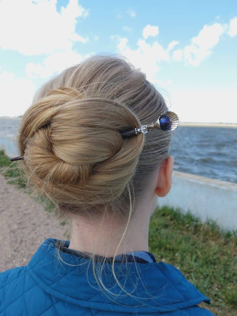 A blonde young lady wearing a dark blue shirt is standing on the sand looking at the sea with long hair twisted in a bun using a Lilla Rose hair stick with a silver sea shell and a royal blue pearl shape inside for the article The importance of taking care of your hair when you are stressed.  