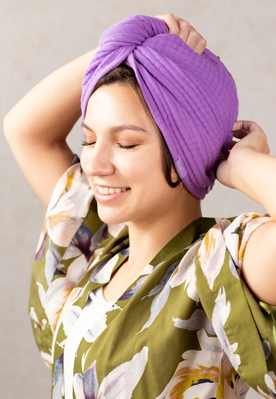 Women in olive green with light colored flowers silk robe securing a purple 100% cotton gauze hair towel from Lilla Rose. Her eyes are closed, and her hands are above her head, one on top and the other near the bottom for the article The importance of taking care of your hair when you are stressed. 