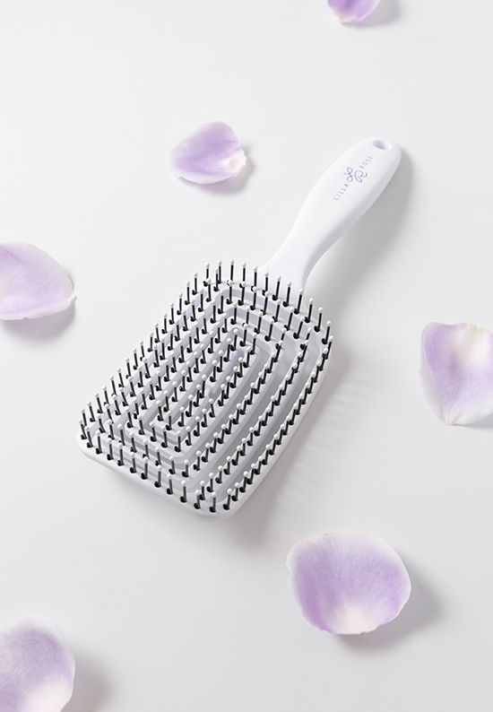 A Lilla Rose Flexi brush has a white handle, a rectangular head, and black bristles on an off-white background surrounded by six light purple petals for the article The importance of taking care of your hair when you are stressed.