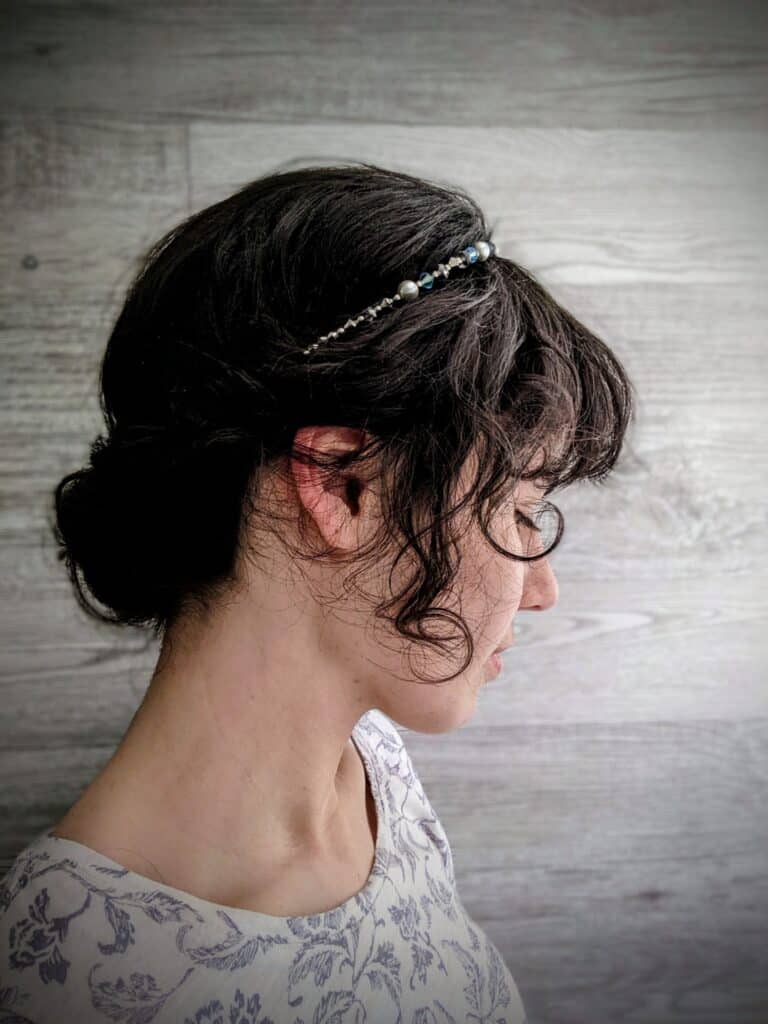 A young lady is standing in front of a pale wood-planked wall. She has dark brown hair rolled into a Lilla Rose hairband in the back. The Lilla Rose hairband is blue, silver, and white. She has brown curls coming down the sides and bangs. She is facing sideways, so we see her profile.  She is under the heading Hairbands for the article Get Your Hair Game On Point with Lilla Rose Accessories - Say Goodbye to Bad Hair Days!