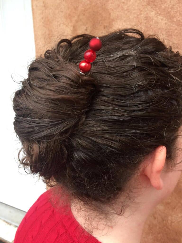 A woman with dark brown hair is twisted in a Lilla Rose stick with three round beads. She is wearing a red shirt and is facing away; we can only see the right rear side exposing the back side of her head and ear. She is Under the heading Hair Sticks in the article Get Your Hair Game On Point with Lilla Rose Accessories - Say Goodbye to Bad Hair Days!