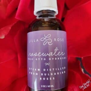 Picture of Lilla Rose Essentials Rose Water in a purple bottle laying on a background of red roses for the article Environmental factors can damage your hair, so keep it protected.