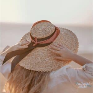 Women wore a woven hat with a ribbon tied around it that is blowing in the wind. She is holding it on with both hands. She is wearing it under the heading Protect your scalp for the article  Why scalp care is important to healthy hair